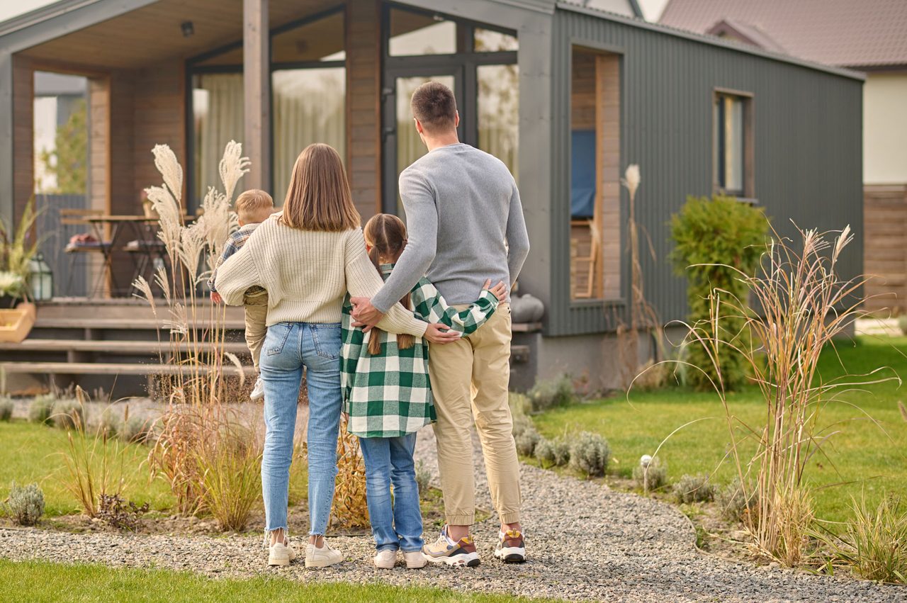 A family of four, seen from behind, looking at their modern home across a manicured lawn.