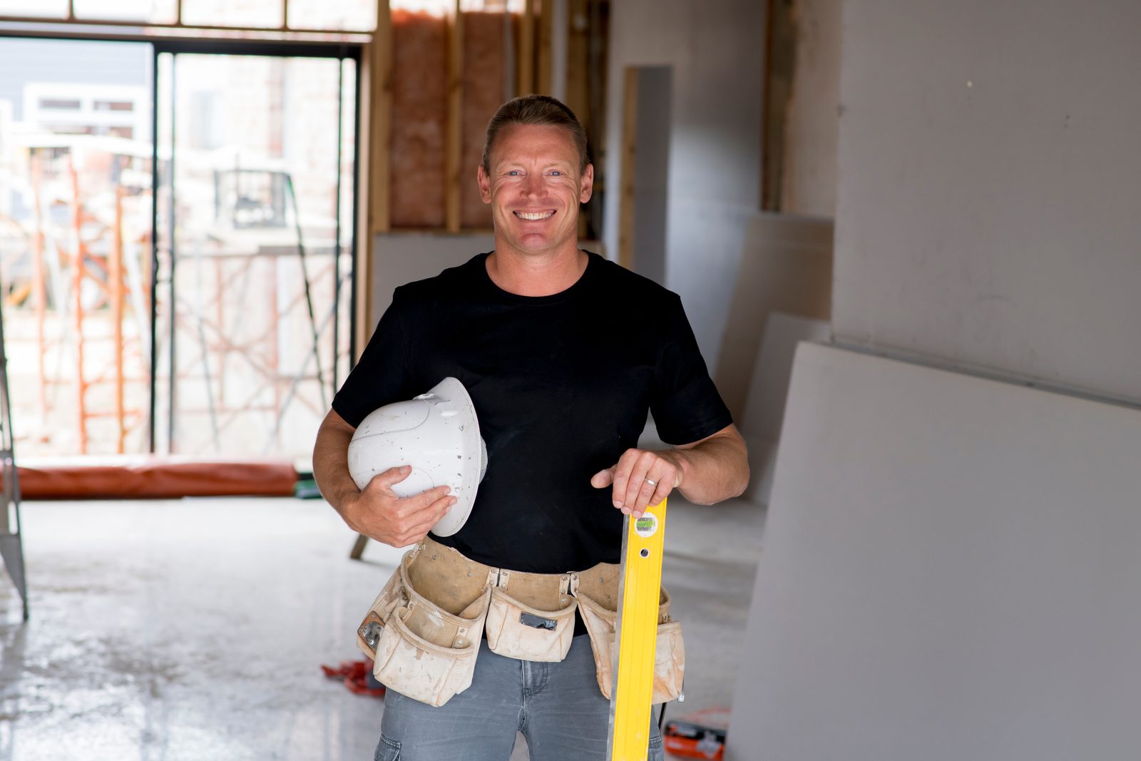 A smiling builder holding a hard hat and a spirit level inside an unfinished building, with site scaffolding visible through the window behind him.
