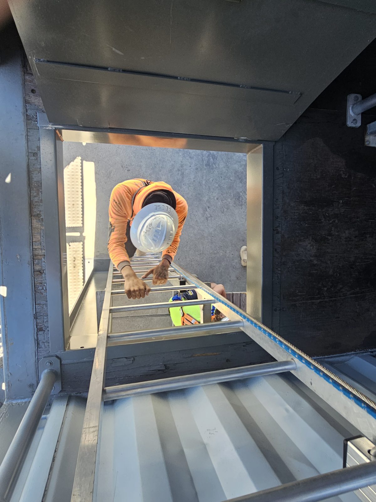 Worker climbing down ladder into narrow vertical confined space wearing hard hat and safety rope