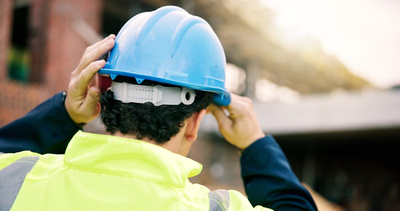 Worker adjusting and securing blue hard hat while wearing high-visibility safety vest and gloves