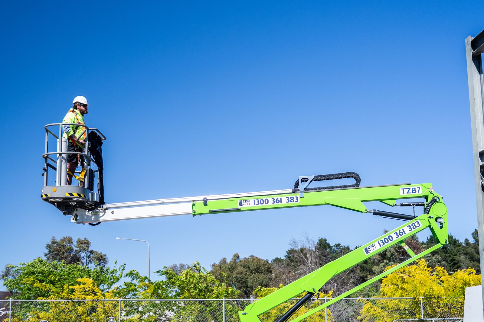 Worker elevated on a green boom lift platform wearing safety harness and high-visibility clothing